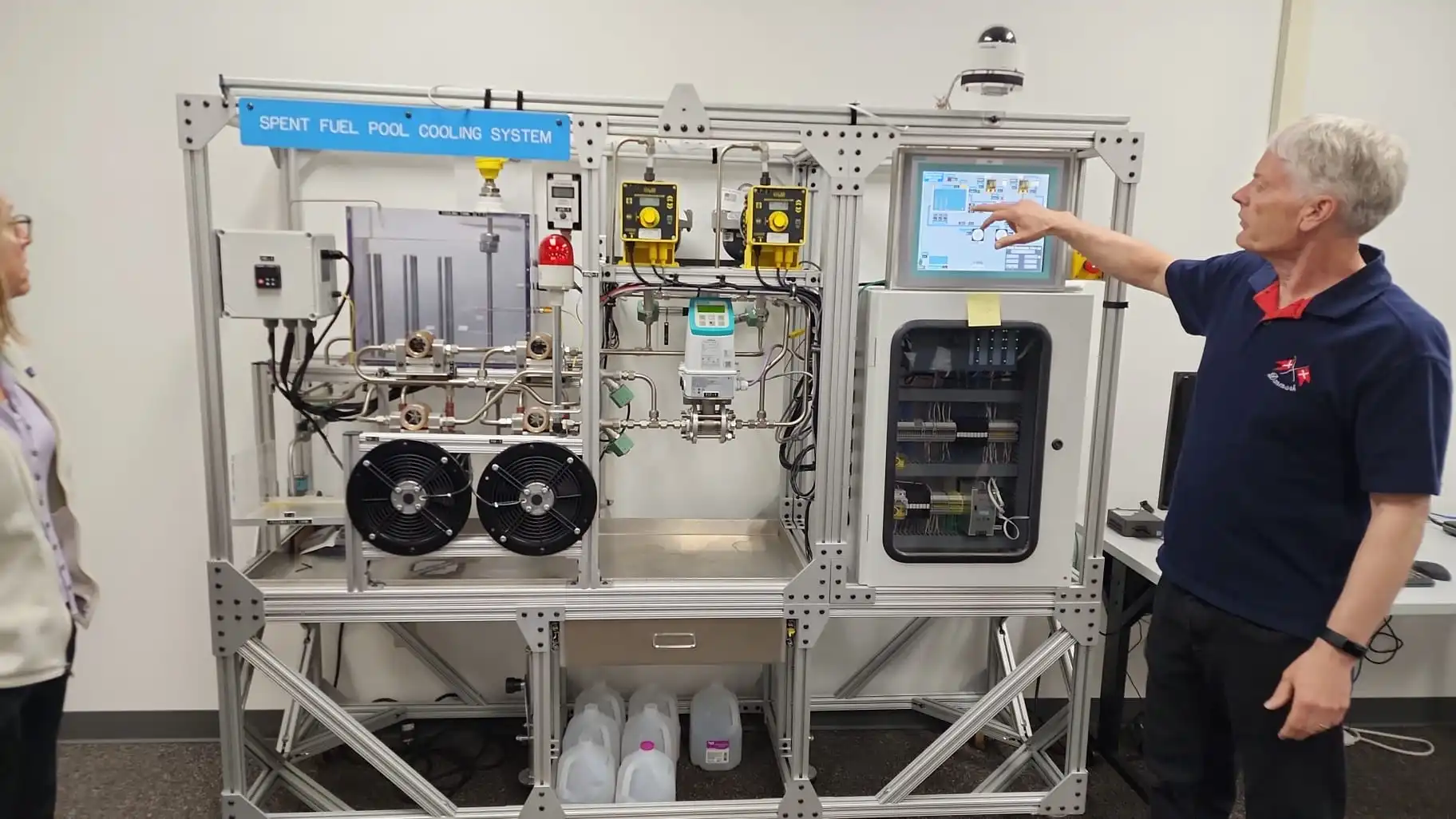 An instructor points to the touchscreen on the Spent Fuel Pool Cooling System training rig, surrounded by pipes, pumps, fans, sensors, and control hardware.