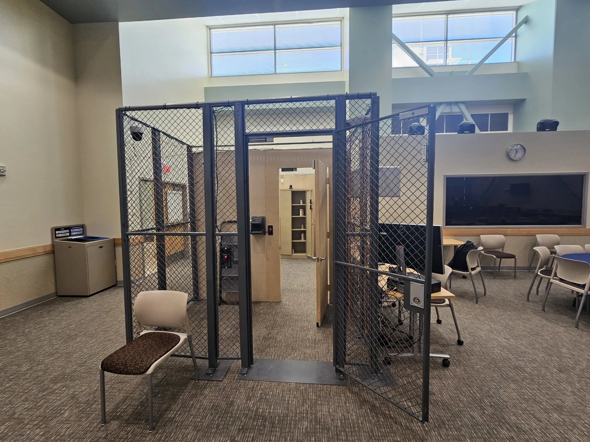 A chain-link security enclosure with an open gate, an interior door, and a secure cabinet with faux-nuclear material stands in a classroom beside workstations and an observation window.